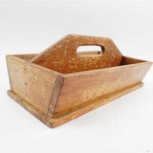 May include: A rectangular wooden caddy with a weathered, light brown finish and a central handle featuring a cutout grip. The storage container has slanted side panels and a flat base, showing visible wood grain and signs of age throughout the surface.