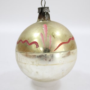 May include: Vintage Christmas ornament with a silver and gold finish. The glass ball is decorated with red wavy lines and pink accents. A metal cap and hanging loop are at the top. A classic holiday decoration.
