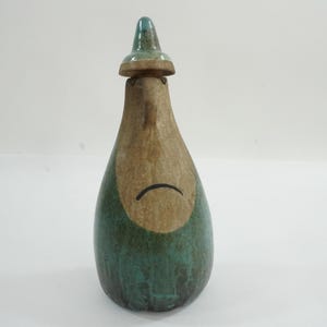 May include: A handcrafted ceramic figurine in the shape of a gnome. The gnome has a brown face and hat, with a teal-colored body. A black curved line represents the mouth. The figurine is approximately 7 inches tall.