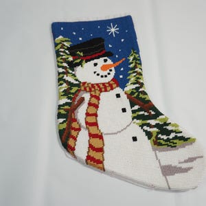May include: Needlepoint Christmas stocking featuring a snowman with a black top hat, orange carrot nose, and a red and gold scarf. The background shows a blue sky with a star and evergreen trees. The stocking is white with a blue top.