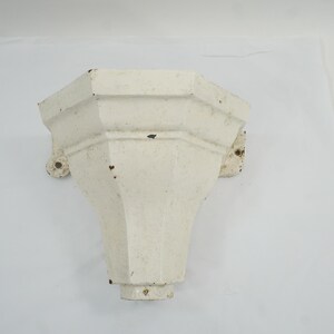 Puede incluir: Un soporte arquitectónico octogonal de hierro fundido blanco con un cuerpo acampanado y cónico, y bridas de montaje visibles en los lados. La superficie presenta un acabado mate desgastado con pequeñas manchas oscuras de desgaste y oxidación.