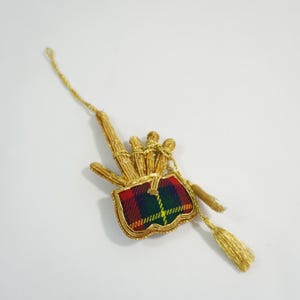 May include: A gold-colored ornament shaped like bagpipes, featuring a red, green, and yellow plaid pattern. The ornament has a gold tassel and a gold cord for hanging. The bagpipe design is detailed with gold thread.