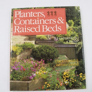 May include: A softcover book titled Planters, Containers & Raised Beds: A Gardener's Guide by Chuck Crandall & Barbara Crandall. The cover features a photograph of a tiered garden with wooden retaining walls, green shrubs, and colorful flowers.