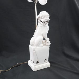 May include: A white ceramic table lamp featuring a sculpted Foo Dog figure perched atop a rectangular pedestal base. The lamp includes a central vertical rod extending upward from the figure, a brown electrical cord, and a plug with a switch.