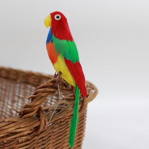 Pode incluir: Uma figurinha de papagaio de penas coloridas, com plumagem vermelha, verde, amarela e azul. O papagaio está empoleirado na borda de uma cesta de vime marrom.