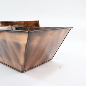 以下が含まれることがあります： 銅色の正方形の金属製容器で、持ち手が付いています。容器は台形型で、上部の開口部が底部よりも広くなっています。金属はわずかに古びた外観をしています。