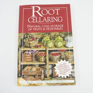 May include: A book titled "Root Cellaring" with the subtitle "Natural Cold Storage of Fruits & Vegetables." The cover features illustrations of a wooden shelf with baskets and crates of produce, including apples, cabbages, and tomatoes. The book also has text that says "Keep your produce harvest fresh in your own basement, porch, garage, or closet hideaway."