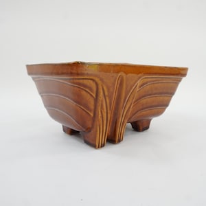 May include: A brown ceramic planter with a square shape and three small feet. The planter has a textured surface with curved lines and a decorative design on the sides.