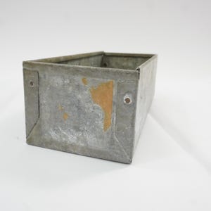 May include: A rectangular galvanized metal bin with a weathered gray finish and visible patches of tan oxidation. The container features reinforced corners, a flat base, and two small circular holes on the front panel for mounting or attachment.