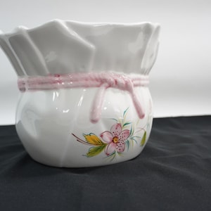 May include: White ceramic vase with a ruffled top and a pink ribbon detail. The vase features hand-painted floral designs in pink, yellow, and green. The vase is a decorative item.