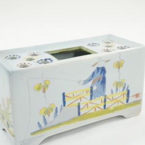 May include: Rectangular ceramic vase with a light blue base, featuring a hand-painted scene of a person fishing. The vase has a square opening and several smaller circular openings on top. Decorative yellow and blue floral accents are also present.