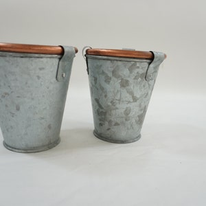 Puede incluir: Dos cubos pequeños de metal galvanizado con asas de color cobre. Los cubos son ligeramente diferentes en tamaño y tienen un acabado desgastado.