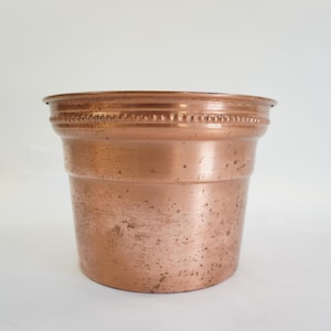 Puede incluir: Una maceta de metal color cobre con forma ligeramente acampanada. El borde presenta un diseño decorativo de cuentas. La maceta tiene un aspecto rústico y envejecido, lo que sugiere que es vintage. Ideal para uso en interiores o exteriores. Mide unos 15 cm de alto.