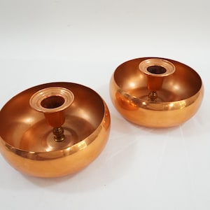 Può includere: Due candelabri in rame con un design a forma di ciotola. Ogni supporto ha una coppa cilindrica centrale per la candela. Il rame ha una finitura calda e riflettente. I candelabri sono posti su una superficie bianca.