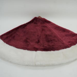 May include: A circular tree skirt featuring a deep burgundy velvet-like fabric center and a wide, plush white faux fur border. The design is semi-circular in shape with a small opening at the top center, showcasing a soft, textured appearance.