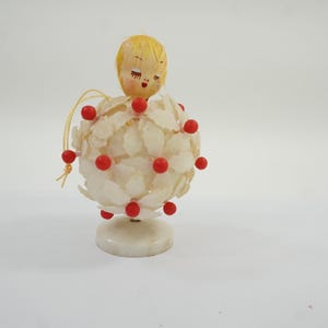 May include: A decorative figurine featuring a round body covered in white, petal-like shapes accented with small red beads. The figure has a painted face with blonde hair and stands on a circular white base, with a thin gold string attached to the side.