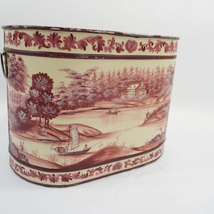 Puede incluir: Una papelera ovalada de metal con fondo color crema y un estampado de paisaje en color rojo oscuro estilo toile. El diseño muestra una escena fluvial con veleros, árboles y una casa, bordeado por un patrón floral decorativo arriba y abajo.