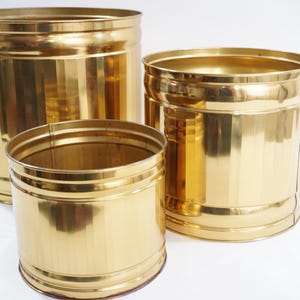Puede incluir: Tres macetas de metal dorado de diferentes tamaños. La más grande es cilíndrica, la del medio es un poco más pequeña y la más pequeña es un cilindro corto y ancho. Todas tienen una superficie brillante y reflectante.