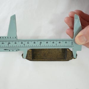 May include: A blue plastic caliper measuring a small, rectangular, gold-colored object. The object is approximately 1.5 inches long.