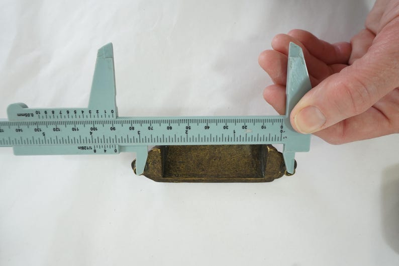 May include: A blue plastic caliper measuring a small, rectangular, gold-colored object. The object is approximately 1.5 inches long.