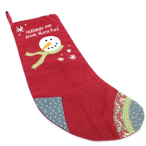 Può includere: Calza di Natale rossa con un pupazzo di neve e fiocchi di neve ricamati. La calza ha un bordo a quadri verdi e bianchi e il testo "Holidays Are Snow Much Fun!" è ricamato sulla calza.