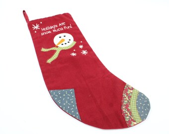 May include: Red Christmas stocking with a snowman and snowflakes embroidered on it. The stocking has a green and white plaid trim and the text "Holidays Are Snow Much Fun!" is embroidered on the stocking.