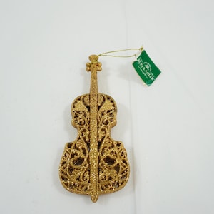 May include: Glittery gold violin-shaped ornament with intricate cut-out details. The ornament has a tag attached with the text "Kurt S. Adler". A festive decoration for music lovers or holiday decor.