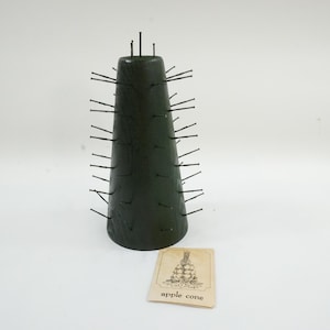May include: A dark green, cone-shaped object with numerous metal pins protruding from its surface. A small card with the words "apple cone" and an illustration of apples is next to the cone. The cone is likely a decorative item.