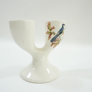 May include: Cream-colored ceramic vase with a unique double-spout design. One side features a detailed illustration of a blue jay perched on a branch with autumn leaves. The vase has a rounded base and a smooth finish.