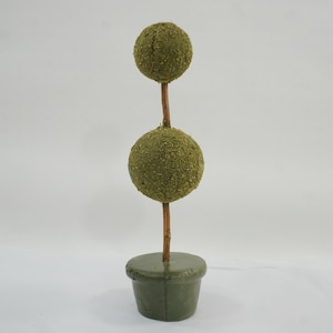 May include: A topiary decoration featuring two stacked green moss-covered spheres on a thin brown stem, supported by a small, round, dark green base. The structure is vertical, with the smaller sphere positioned above the larger sphere.