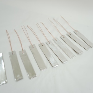 May include: A collection of silver-colored metal bookmarks with a rectangular shape and a small hole at the top. Each bookmark has a thin, copper-colored string attached. The bookmarks are arranged diagonally on a white surface.