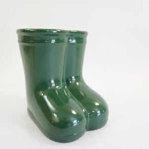 May include: A glossy, dark green ceramic rain boot planter. The planter is shaped like two boots side-by-side, with a rounded toe and a smooth, reflective surface. The boots are approximately 20 cm tall.