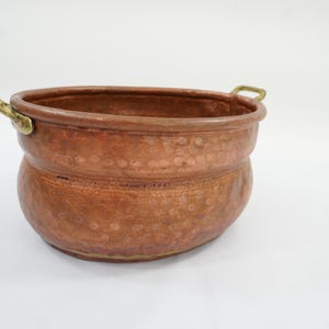 May include: A round, hammered copper pot featuring a wide, curved body and a smooth, reinforced rim. The vessel includes two small, ornate brass handles attached to opposite sides of the upper edge, providing a contrast to the warm, metallic copper finish.