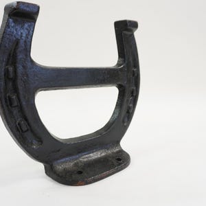 May include: A cast iron horseshoe-shaped wall hook in a dark, weathered finish. The hook has a flat base with two holes for mounting and a curved top with visible nail holes, suggesting a rustic or vintage aesthetic.