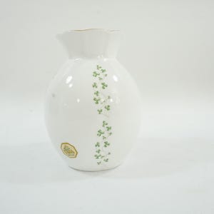 May include: White ceramic vase with a ruffled rim and a gold trim. The vase features a vertical design of green shamrocks. A gold and white sticker is on the lower part of the vase.