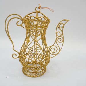 Peut inclure: Une théière décorative en fil de fer doré et pailleté, dotée d'un motif en filigrane ajouré. L'objet présente une anse courbée, un long bec verseur et une petite boucle sur le dessus pour la suspension, formée de fils métalliques entrelacés.
