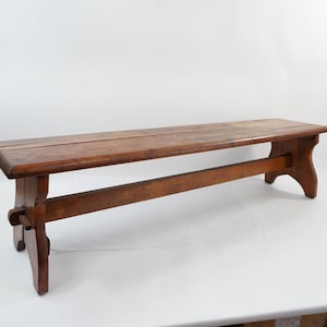 May include: A medium-brown wooden bench featuring a rectangular seat with rounded edges, supported by two side legs connected by a horizontal stretcher bar. The wood grain is visible throughout the surface, showing a natural finish.