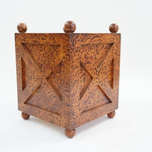 May include: A square, wooden planter with a textured, speckled finish in shades of brown. The planter features geometric cutouts and decorative ball feet and top accents. The design is symmetrical and the overall aesthetic is rustic.