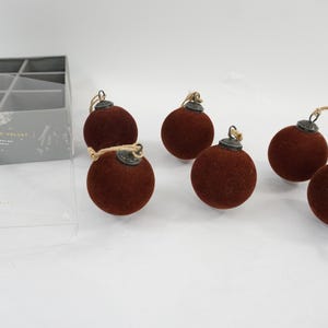 May include: A set of six spherical ornaments covered in dark brown flocked velvet, each featuring a metallic cap and a jute twine loop for hanging. The ornaments are displayed alongside a gray storage box with a clear lid labeled Flocked Velvet.