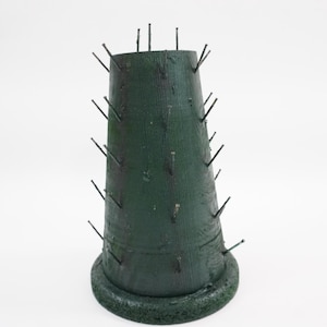 May include: A dark green, cone-shaped floral foam base featuring a textured surface and a circular bottom rim. Numerous thin, dark metal pins are inserted at regular intervals around the exterior of the cone, protruding outward from the foam.