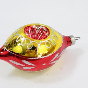May include: A vintage Christmas ornament with a teardrop shape. The ornament is red with silver glitter accents and a yellow and red top. The top has a decorative design. A silver metal loop is attached for hanging.