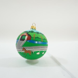May include: A green glass Christmas ornament with a painted scene of a red barn, a bridge, a horse, and sheep. The ornament is on a white background.
