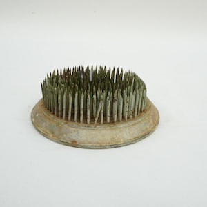 Op de afbeelding: Een vintage metalen bloemenkikker met een cirkelvormige basis en talrijke scherpe, rechtopstaande pinnen. De pinnen zijn groen getint en de basis heeft een verweerde, verouderde uitstraling. Deze bloemschikkersgereedschap is ontworpen om bloemstelen op hun plaats te houden.