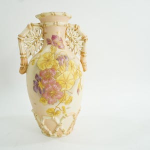 Puede incluir: Un jarrón de porcelana blanca con diseños florales en tonos rosa, amarillo y morado. El jarrón tiene asas esculpidas intrincadas que se asemejan al bambú.
