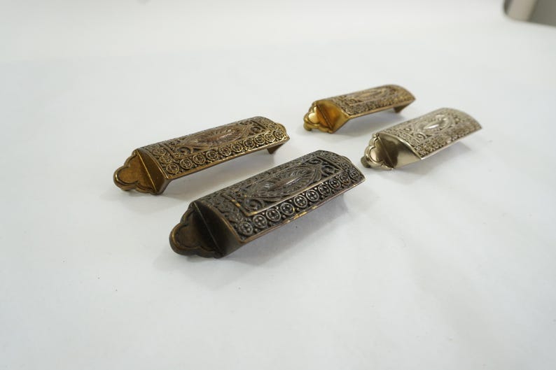 May include: Four ornate metal drawer pulls with an intricate design. The pulls are in different finishes: gold, silver, and black.