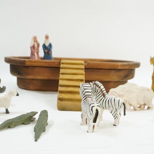 Könnte beinhalten: Ein hölzernes Arche Noah-Set mit verschiedenen Tierfiguren, darunter Elefanten, Zebras, Giraffen und Pinguine. Die Arche ist eine braune Holzschale mit einer Rampe. Die Tiere sind in verschiedenen Farben bemalt.