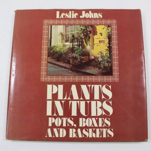 Puede incluir: Un libro rojo titulado "Plants in Tubs, Pots, Boxes and Baskets" de Leslie Johns. La portada muestra una foto de un edificio de ladrillo con una puerta amarilla y una variedad de plantas en macetas y cubetas.