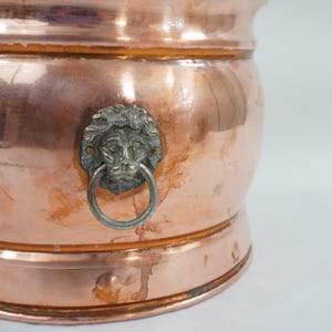 May include: A polished copper vessel featuring a rounded body and a prominent silver-toned metal lion head ornament. The lion head is centered on the exterior and holds a circular ring in its mouth, serving as a decorative handle detail.