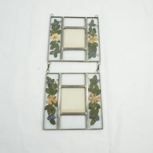 May include: Two rectangular glass photo frames connected by a chain. Each frame has a central square for a photo, surrounded by clear glass panels. The frames are decorated with pressed flowers in yellow, blue, and green.