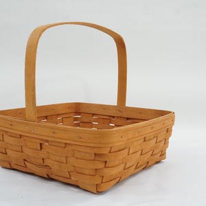 May include: A square-shaped woven basket made of light brown wood strips. The basket features a single tall, arched handle attached to the centre of two opposite sides. The rim is reinforced with a smooth wooden band secured by small metal nails.